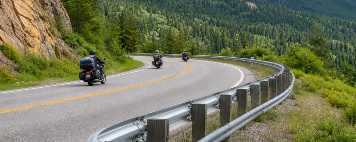 Motorcycles traveling on a mountain road beside a guardrail with a lower auxiliary rail