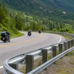 Motorcycles traveling on a mountain road beside a guardrail with a lower auxiliary rail